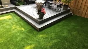Backyard with artificial grass and raised deck featuring potted plants and outdoor furniture.